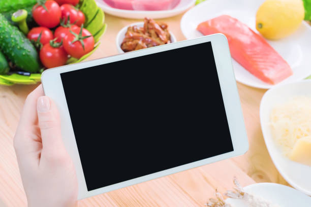 istock Close-up top view of female hands holding a tablet computer with a blank display surrounded by healthy food on a home kitchen table. Workpiece to insert the design of the mobile application into the screen. Products of Greek cuisine raw seafood and fresh 949623848