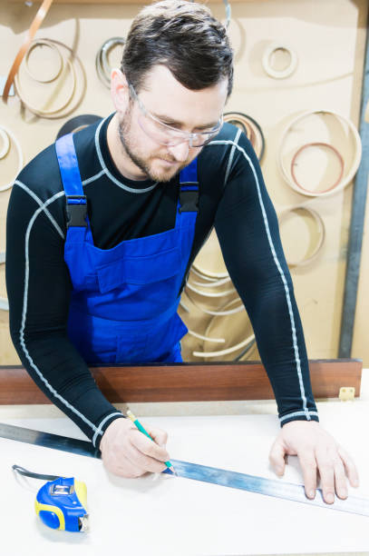 A bearded tired handsome carpenter on a table with a pencil draws a sign on the board. At the workplace there is an ordinary master dressed in blue cambenizon with a cruel haircut and saved glasses. The concept of garage furniture production. un barbudo cansado carpintero guapo en una mesa con un lápiz dibuja una señal en el tablero. en el lugar de trabajo hay un maestro ordinario de vestida de azul cambenizon con un corte de pelo cruel y guarda gafas. el concepto de producción de muebles de la cochera