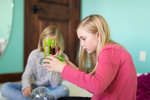 Two blonde elementary-age girls play with a science kit on floor. The kit consists of two separate plastic molded pieces for studying physics and electricity. The girl in the foreground is lifting it up carefully and examining the amount of liquid in the beaker.