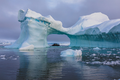 A zodiac full of tourist viewed through an arch in a large blue iceberg with reflections, Antarctica