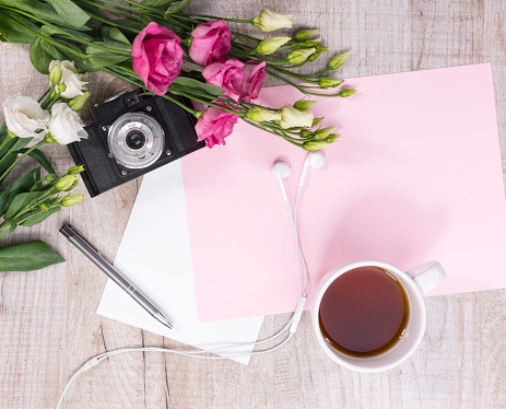 Top view of cup of tea, flowers, vintage camera, earphones, pen and sheet of paper on a light wooden background. Flat lay style