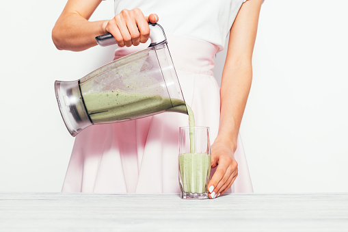 Young woman fills glass with fresh made spinach-banana smoothie on white background. Simple and elegant image with copy space.