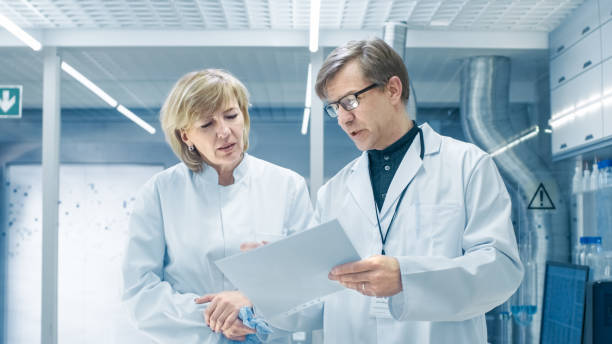 mujeres y hombres científicos jefe caminando a través de modernos laboratorios y discutiendo las posibilidades futuras. joven ayudante trabaja en su escritorio. - scientist lab coat doctor male fotografías e imágenes de stock