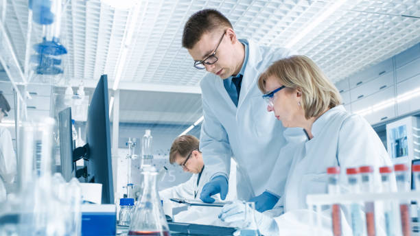 en moderno laboratorio científico femenino tiene discusión con auxiliar de laboratorio masculino joven. muestra sus tablas de datos en un portapapeles, analiza y entra en su ordenador. - scientist lab coat doctor male fotografías e imágenes de stock