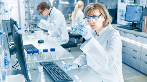 istock Female Scientist Analyzes Liquid in the Beaker and Types Down Observations on Her Computer. She's Working in a Busy Laboratory Full of Scientists Conducting Experiments. 949947072