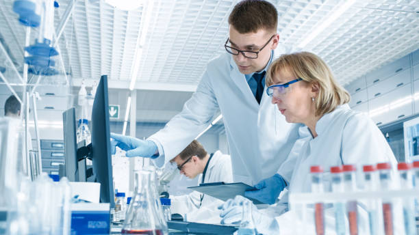 dans le laboratoire moderne scientifique féminin a discussion avec jeune laborantine mâle. il montre ses tableaux de données dans un presse-papiers, elle analyse il et il pénètre dans son ordinateur. - scientist research group of people analyzing photos et images de collection