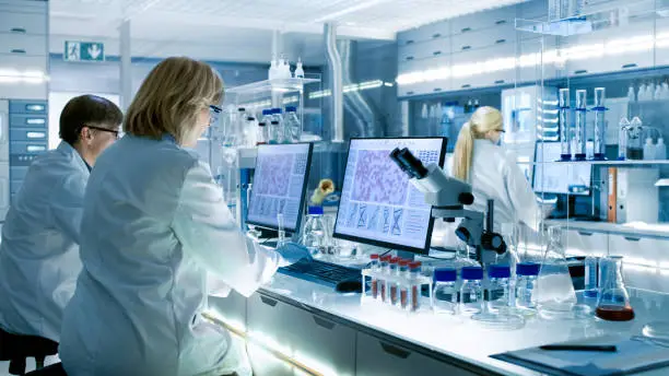 Photo of Female and Male Scientists Working on their Computers In Big Modern Laboratory. Various Shelves with Beakers, Chemicals and Different Technical Equipment is Visible.