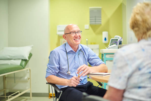 friendly senior gp senior male gp at his desk chatting with a patient general practitioner stock pictures, royalty-free photos & images