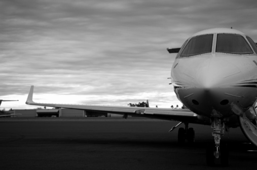 this is a black and white picture of a private luxury Jet at a airport. the jet or plane has it's landing gear and the jet is waiting to be boarded by passengers. the photo shows the jet's or plane's wing, pilot's cabin. in the background of the photo there are airplane hangers that house these private jets and commercial jets and planes. 