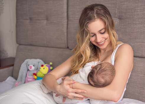 Breastfeeding baby. Pretty mother holding her newborn child. Mom smile and nursing infant. Beautiful woman and new born love at home. Blond mother breast feeding baby.