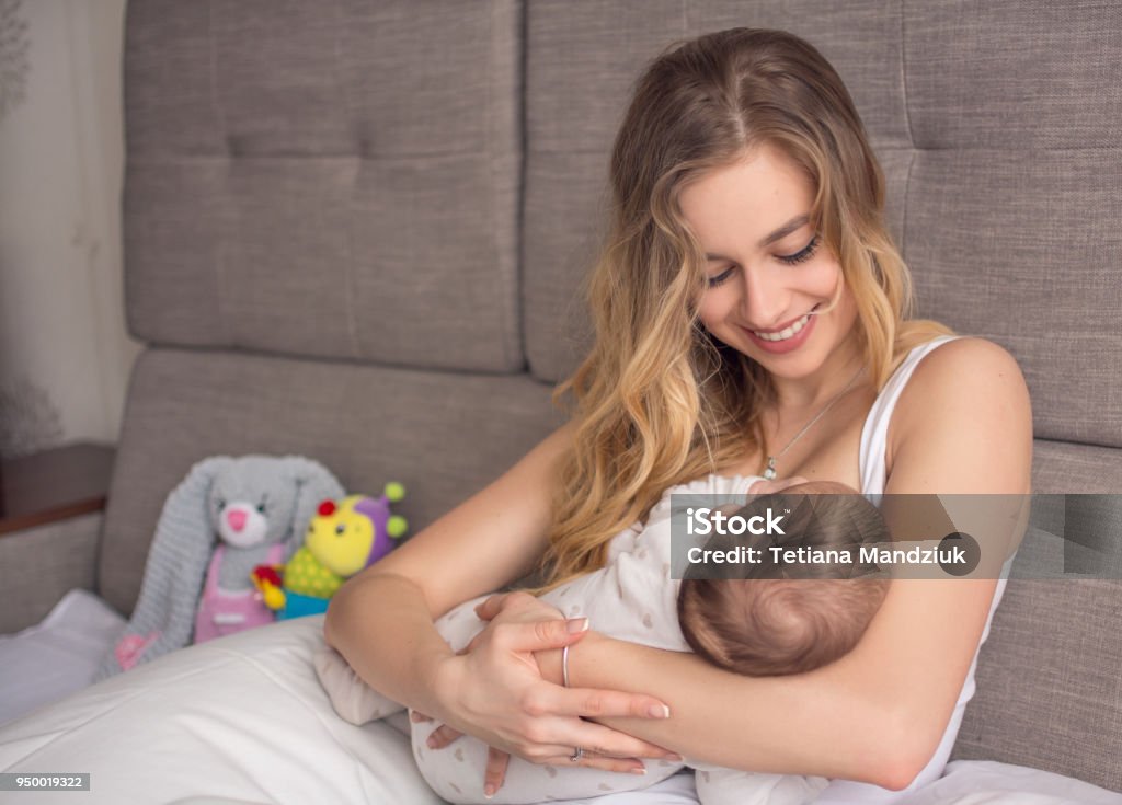 Breastfeeding baby. Pretty mother holding her newborn child. Mom smile and nursing infant. Beautiful woman and new born love at home. Blond mother breast feeding baby. Breastfeeding Stock Photo