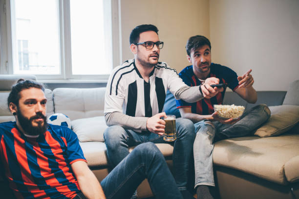 istock Friends in soccer jerseys using remote control while watching TV at home 950020534