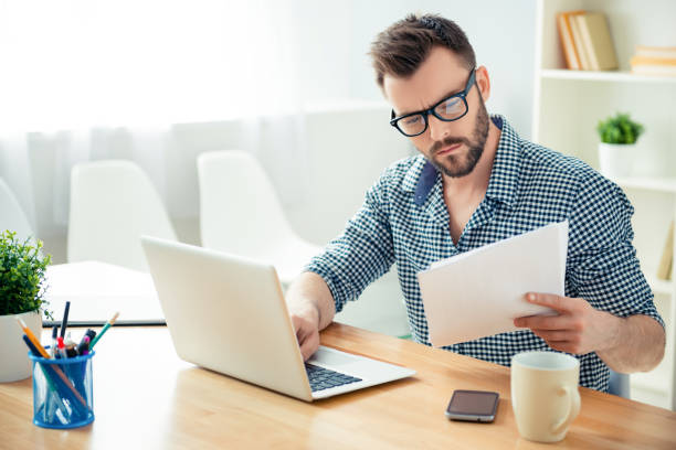 portrait of concentrated businessman in glasses with laptop reading contract - paper document notebook laptop imagens e fotografias de stock