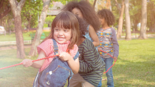 kid girls and boys pulling a long red rope, playing tug of war in the park that look like they are very fun and happy to play with friends - tug o war imagens e fotografias de stock