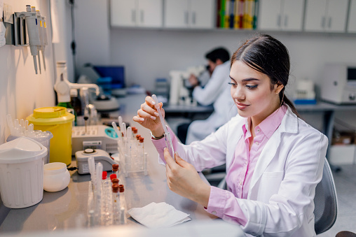 Closeup portrait of young scientist holding conical tube with blue liquid solution, laboratory experiments, isolated lab background. Forensics, genetics, microbiology, biochemistry. Photo of Female medical or research scientist or doctor looking at a test tube of clear solution in a lab or laboratory with her college in background.