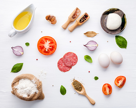 The ingredients for homemade pizza with ingredients sweet basil ,tomato ,garlic ,bay leaves ,pepper ,onion and mozzarella cheese on white wooden background with flat lay.