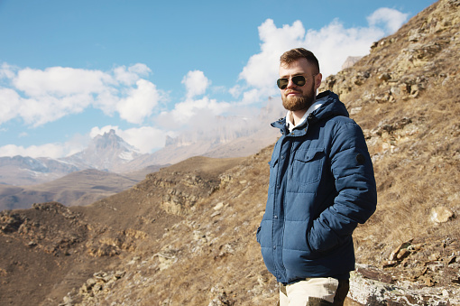 Hipster traveler in a down jacket and sunglasses stands on a mountain slope against the backdrop of epic rocks and smiles. The concept of finding happiness in the mountains.