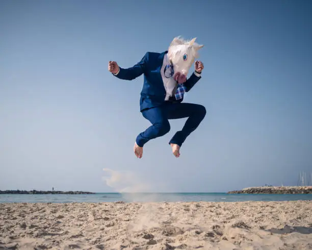 Stylish man in funny mask and suit jumps on beach. Unicorn have fun on the shore and enjoys vacation. Elegant traveler on background of sea and sky