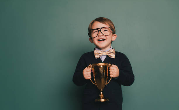 Young Nerd Boy Holding First Place Trophy A young nerd boy wearing a bow tie and eyeglasses with a cheesy smile holds the winning contest trophy. He standing in class in front of a blackboard with his spelling bee trophy. He loves education and loves being smart. spelling bee stock pictures, royalty-free photos & images