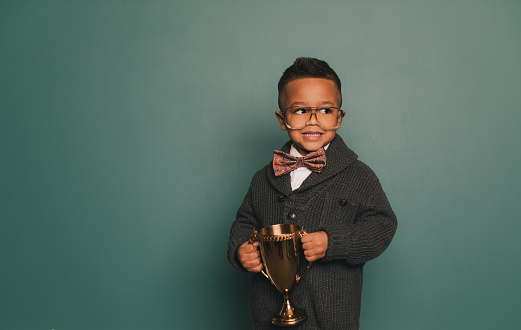 A young nerd boy wearing a bow tie and eyeglasses with a cheesy smile holds the winning contest trophy. He standing in class in front of a blackboard with his science fair trophy. He loves education and loves being smart.