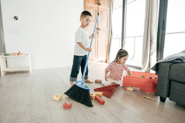 A boy is sweeping the floor while his sister is putting thr toys inside the box.