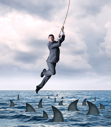 A businessman hangs on for dear life from a rope as he swings above a group of circling sharks in the ocean.  He looks down as the fear from falling into the dangerous waters grips his face.