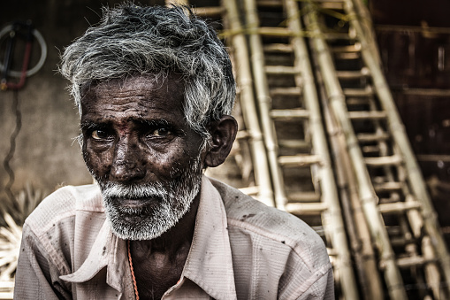 Portrait of Indian elder man with traditional bindi as a third eye, white beard and bamboo ladders on the background in Mysore, Karnataka, India. Concerned expression