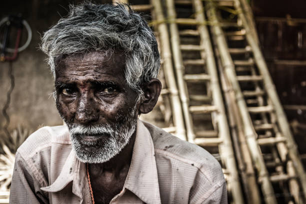 Rural village dark skin Indian old man Portrait of Indian elder man with traditional bindi as a third eye, white beard and bamboo ladders on the background in Mysore, Karnataka, India. Concerned expression india poverty stock pictures, royalty-free photos & images