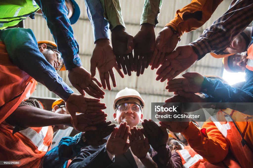Coming together in a circle for unity Owner looking down towards the camera in the center, with all his factory employees huddled up in a circle with their arms in the center. Teamwork Stock Photo