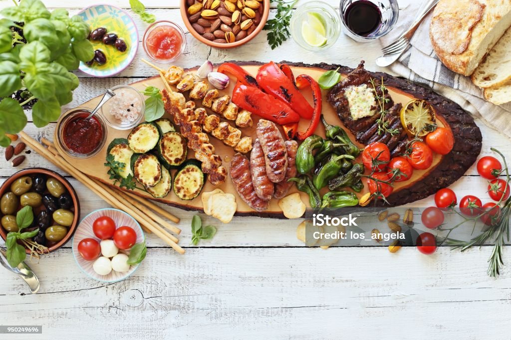 Grilled meat, chicken skewers and sausage  with roasted vegetables and appetizers variety serving on party outdoor table. Mediterranean dinner table concept. Grilled meat, chicken skewers and sausage  with roasted vegetables and appetizers variety serving on party outdoor table. Mediterranean dinner table concept. Overhead view. Barbecue - Meal Stock Photo