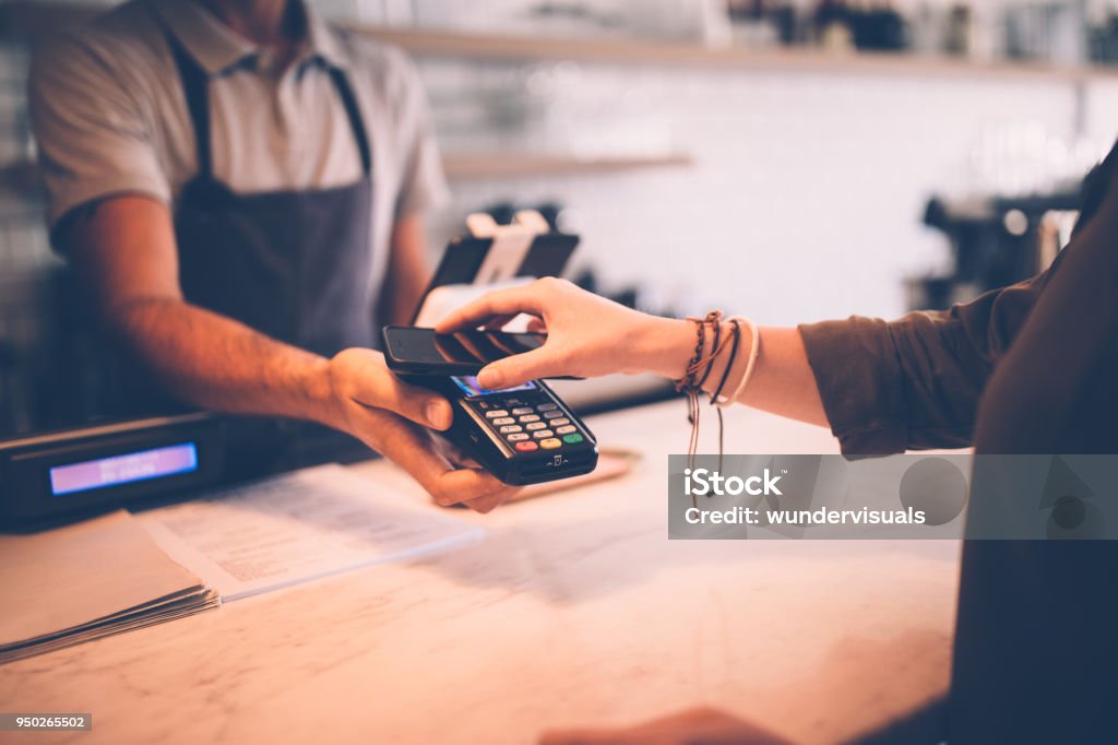 Young hipster woman paying using NFC smartphone contactless technology Young hipster woman paying at restaurant counter using mobile phone contactless NFC technology Mobile Payment Stock Photo