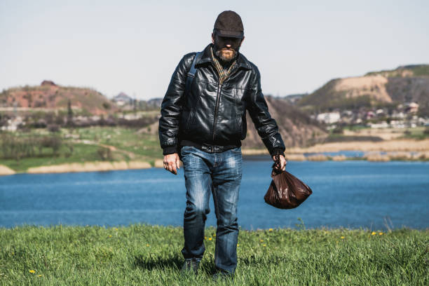 Stylish man in glasses, brown jacket and hat with bag posed on green field.