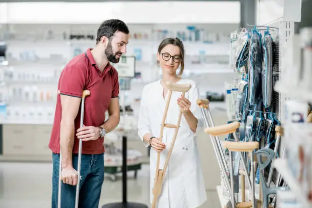 Young woman pharmacist helping to choose crutches for the man in the pharmacy supermarket