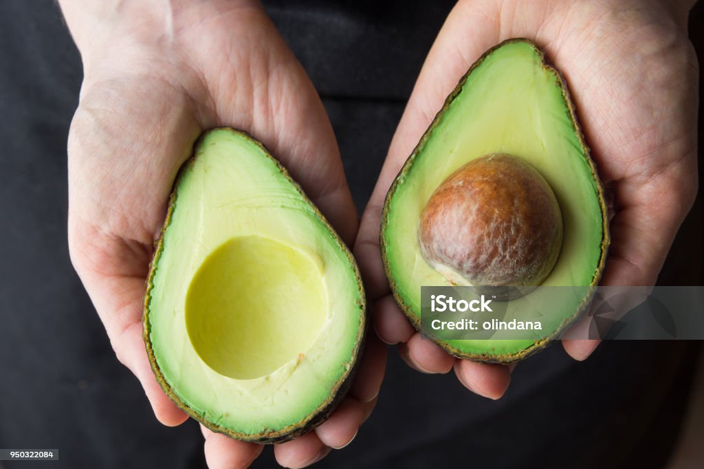 Young Caucasian Woman in Black Apron Holding in Hands Ripe Beautiful Halved Organic Australian Avocado with Pit. Healthy Lifestyle Vegan Vitamin E Oil. Creative Minimalist Food Poster Banner. Kinfolk Young Caucasian Woman in Black Apron Holding in Hands Ripe Beautiful Halved Organic Australian Avocado with Pit. Healthy Lifestyle Vegan Vitamin E Oil. Creative Minimalist Food Poster Banner Avocado Stock Photo