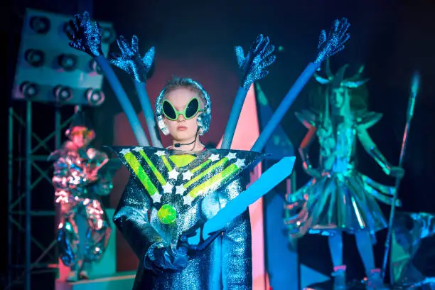 Three children dressed in silver foil space costumes. The children elementary ages are posing together. The serious boy in the space alien costume is standing on foreground. All children are looking at the camera. Studio shooting on black background with a spaceship