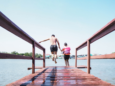 Back view of Asian man and his sister jumping off wooden bridge into river over fisherman village background. Family summer vacation lifestyle at Homestay in Thailand.