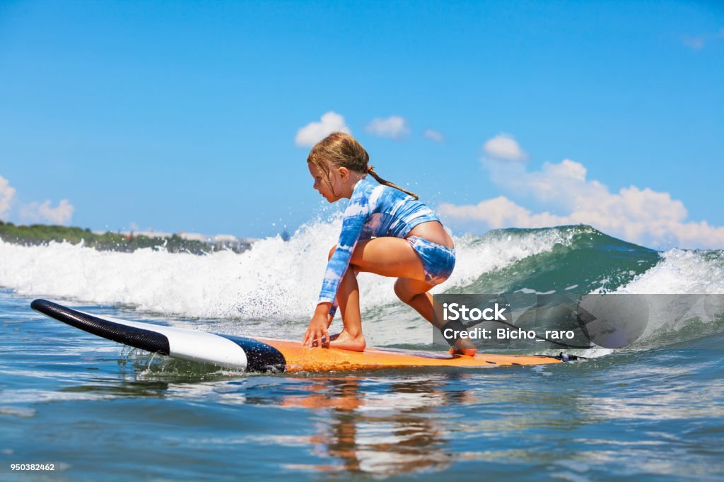 Young surfer rides on surfboard with fun on sea waves Happy baby girl - young surfer ride on surfboard with fun on sea waves. Active family lifestyle, kids outdoor water sport lessons and swimming activity in surf camp. Summer vacation with child. Surfing Stock Photo