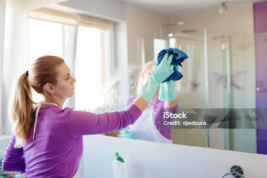 Young beautiful woman in cleaning mirror in bathroom Young beautiful woman cleaning mirror in bathroom. Cleaning service. Maid cleaning at home Cleaning Stock Photo