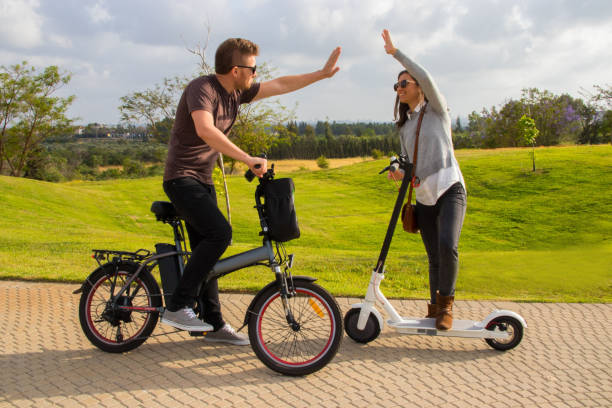 a smiling young  man and a woman with electric bicycles and an electric scooter give high five for each other in the park - two wheel imagens e fotografias de stock