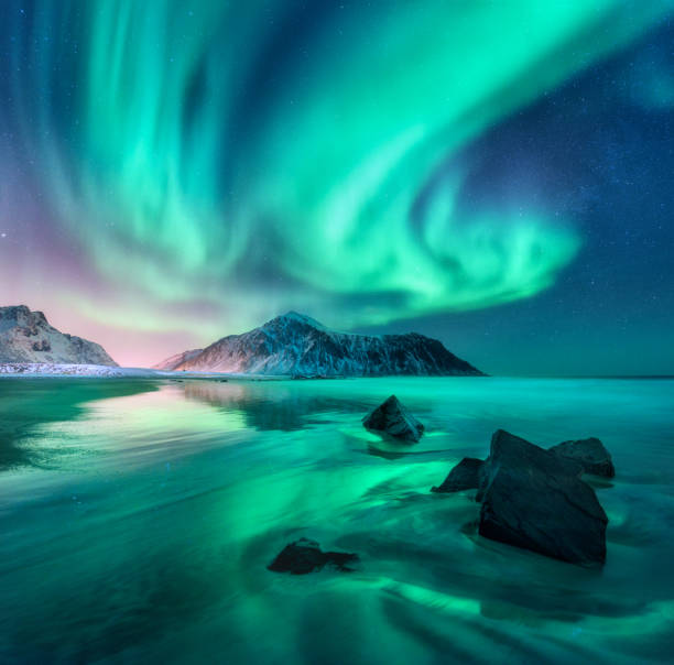 Photo of Aurora. Northern lights in Lofoten islands, Norway. Sky with polar lights, stars. Night winter landscape with aurora, sea with sky reflection, stones, sandy beach and mountains. Green aurora borealis