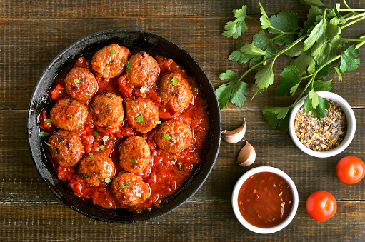 Meatballs in cast iron pan, fresh parsley and tomatoes on wooden table, top view