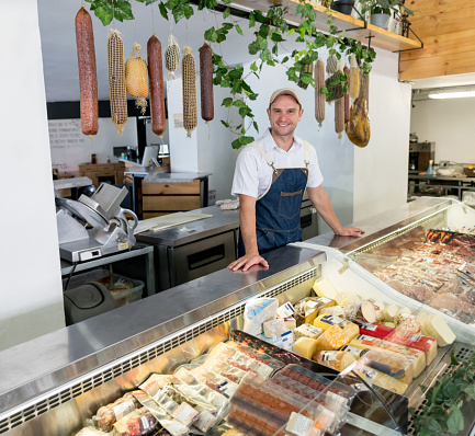 Happy man working at a delicatessen selling meat and cheeses while looking at the camera smiling - supermarket concepts
