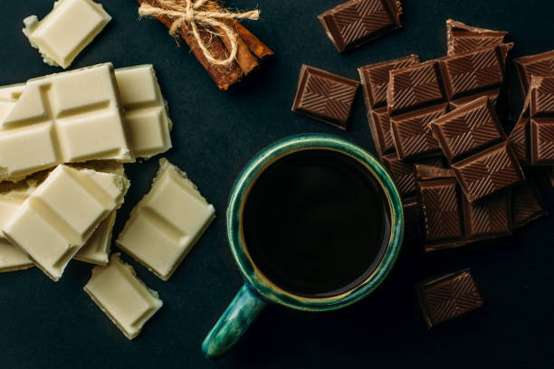 istock Cup of coffee, broken white and black chocolate bars and stack of cinnamon on black table, top view. Tasty energy calorie food 950516716
