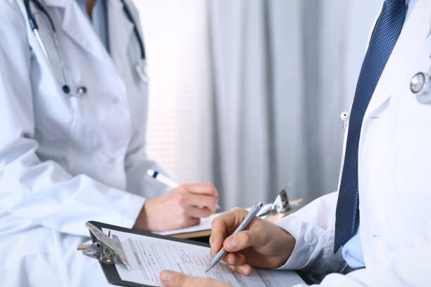 istock Two unknown doctors filling up medical form on clipboard, just hands closeup. Physicians asking question to patient or discussing medication program. Healthcare, insurance and medicine concept 950572480