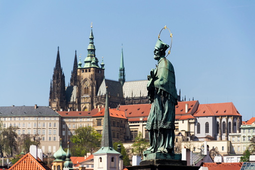 Statue of Saint John of Nepomuk holding crucifix with Jesus Christ on Charles Bridge with Prague Castle and St. Vitus Cathedral in background, Prague, Czech Republic, sunny day, clear sky