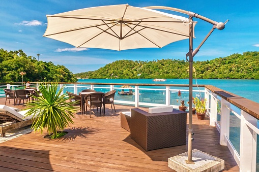 Vibrant blue sky, turquoise water and green trees in the background. The deck is made of natural materials, wood and stone. There are tables and chairs and a sun umbrella provides shade.