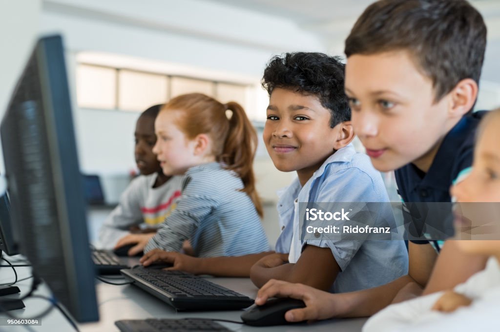 Children using computer in school Multiethnic school kids using computer in classroom at elementary school. Portrait of arab boy looking at camera in a computer room. Smiling primary student in a row using desktop pc in class room. Child Stock Photo