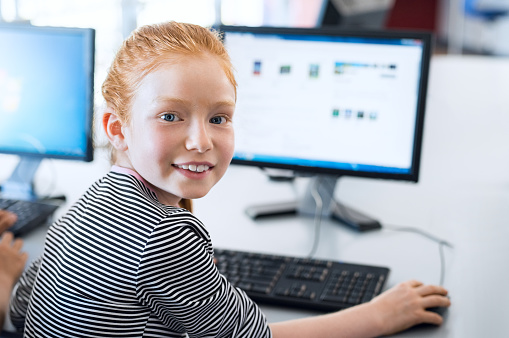 Young girl with red hair using computer at elementary school. Happy female child learning to use internet in a computer room. Portrait of young student looking at camera while typing on keyboard.