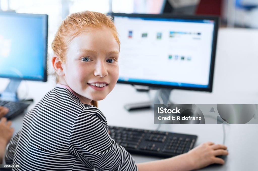 Pupil girl using computer at school Young girl with red hair using computer at elementary school. Happy female child learning to use internet in a computer room. Portrait of young student looking at camera while typing on keyboard. Child Stock Photo