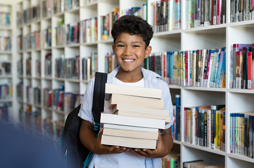 Middle eastern boy holding a stack of books against multi colored bookshelf in library. Portrait of happy arab schoolboy with bagpack holding a heap of books at school. Happy young indian child holding books.